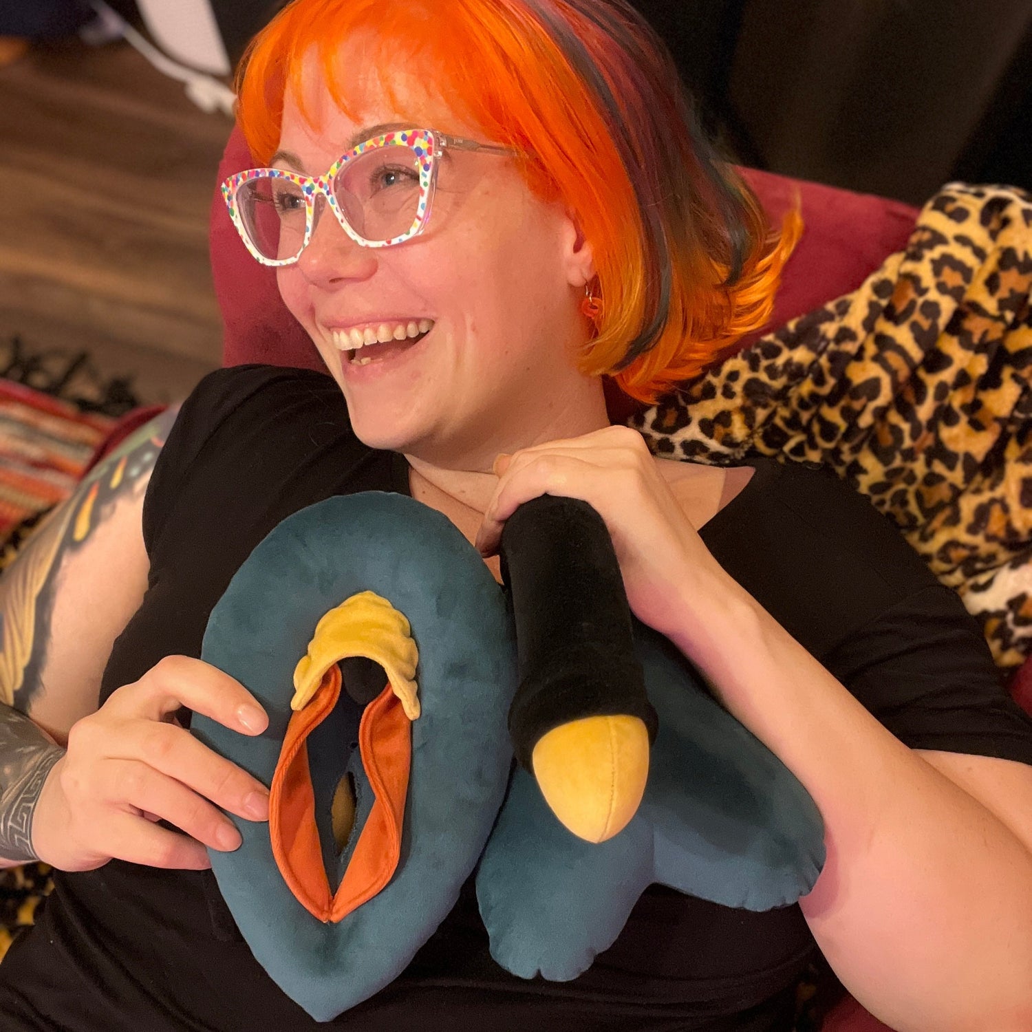 Smiling person with orange hair and polka dot glasses holding a stuffed model vulva and penis